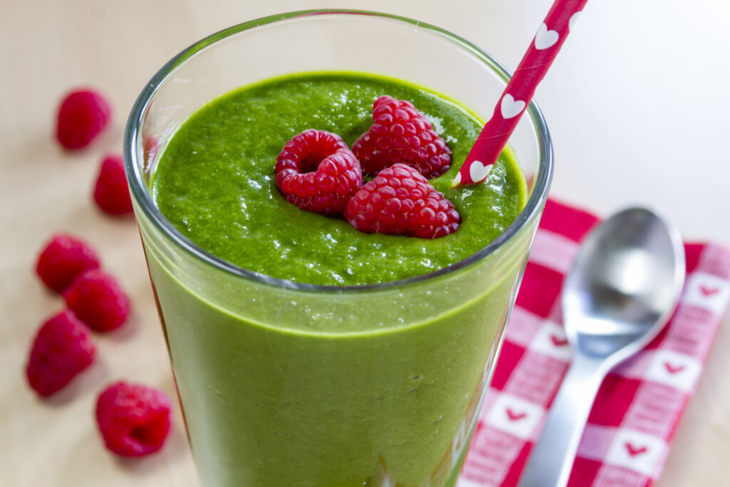 Copo de vidro com suco verde e três framboesas em cima e canudo vermelho com corações brancos