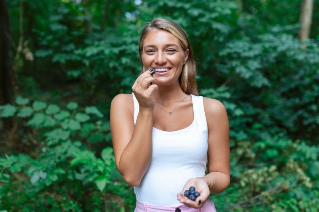 Mulher vestindo blusa branca em pé em um jardim comendo mirtilo