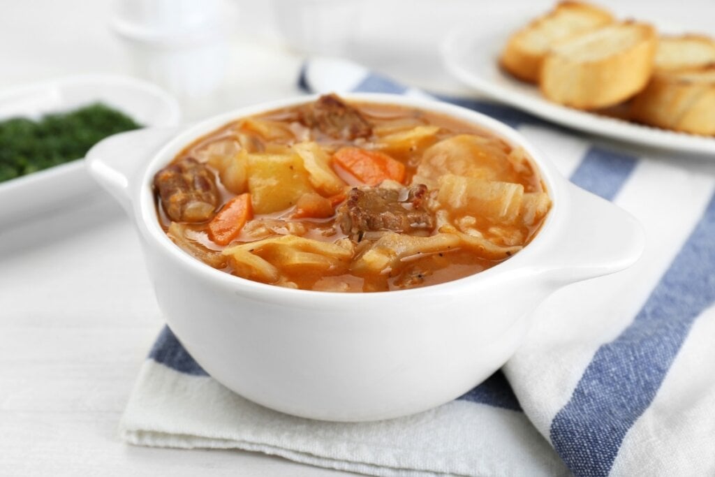 Recipiente branco com sopa de músculo com legumes e macarrão em cima de uma mesa de marmore branco