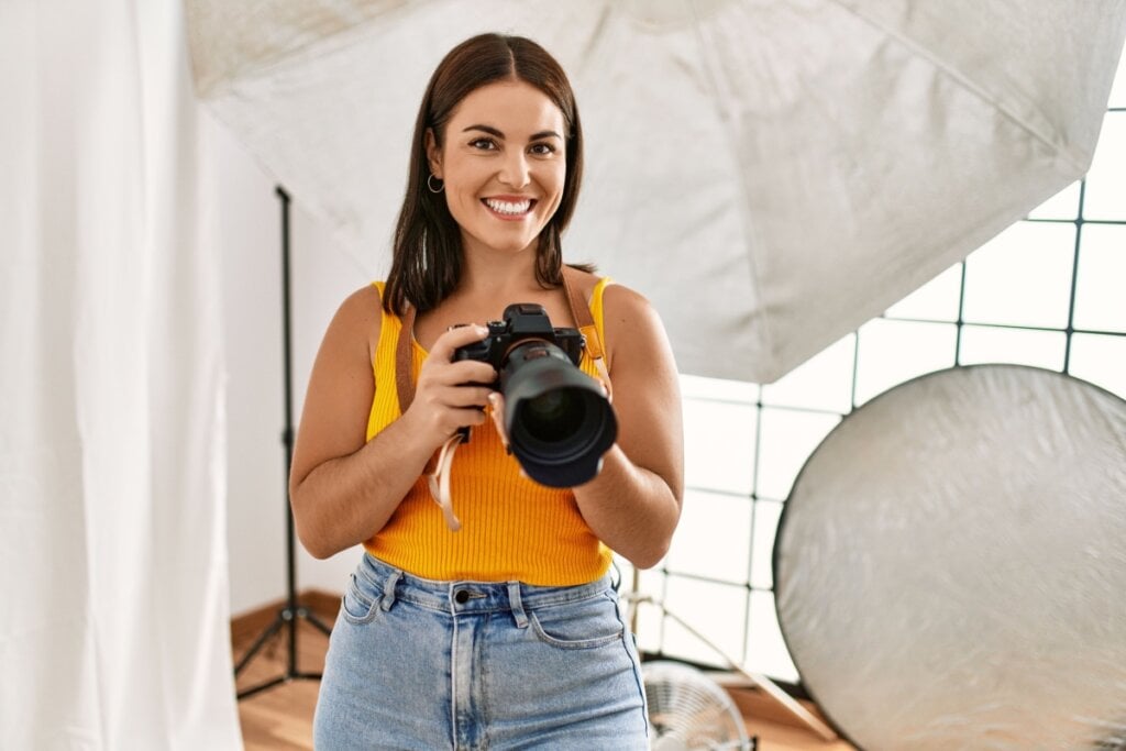 Fotógrafa segurando câmera e sorrindo; atrás há equipamentos de iluminação