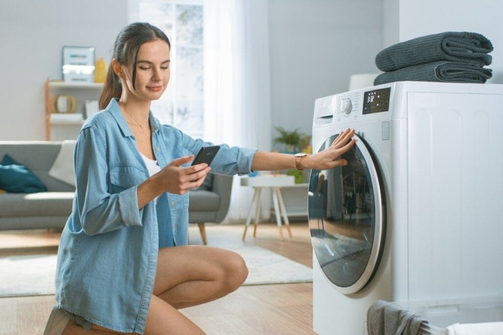 Mulher olhando o celular com a mão em frente a uma máquina de lavar roupa