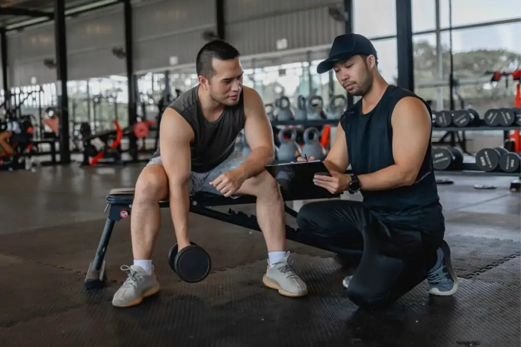 Homem sentado e segurando um peso com um personal trainer do lado 