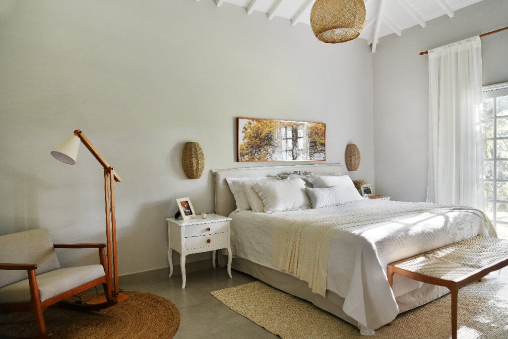 quarto com decoração em tons de branco e bege. cama grande branca. com a madeira na estrutura da poltrona, da luminária de piso e do banco aos pés da cama (com assento palhinha)