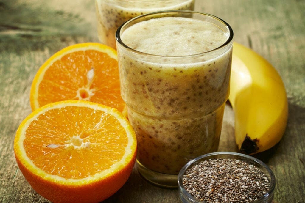 Copo de vidro com suco amarelo em cima de uma mesa de madeira com rodelas de laranja e uma banana