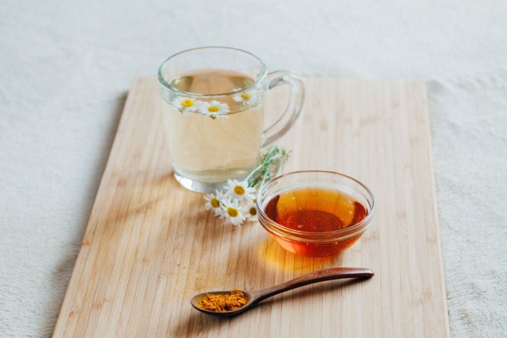 Imagem de caneca de vidro com chá com folhas de camomila. ao lado da caneca, pote com mel e colher com cúrcuma