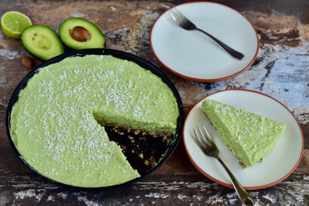 torta de abacate com um pedaço cortado em prato ao lado e um abacate cortado ao meio do outro lado e um prato vazio com um garfo 