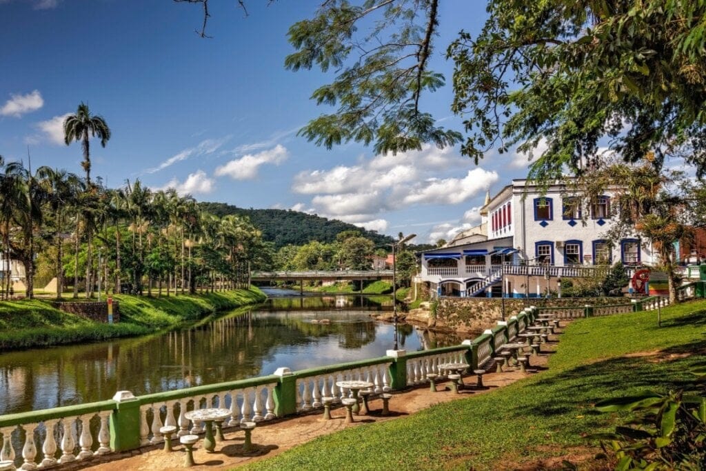 Imagem de um lago com mesinhas e banquinhos de pedra ao redor, ao fundo um prédio histórico em branco e azul, cercado por arvores