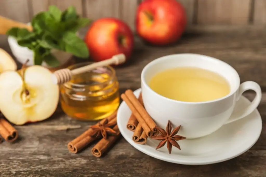 xícara branca com chá amarelo em cima de pires branco com canela e anis-estrelado. maçãs e pote com mel ao fundo