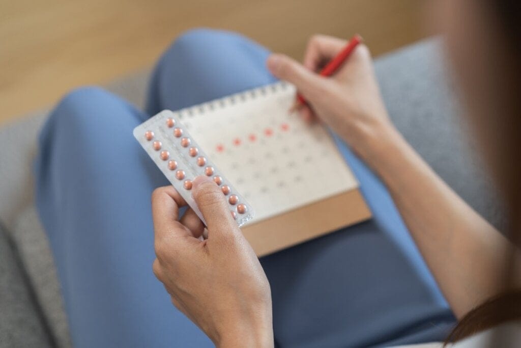 Mãos de uma mulher segurando uma cartela com anticoncepcional e riscando um calendário com uma caneta vermelha