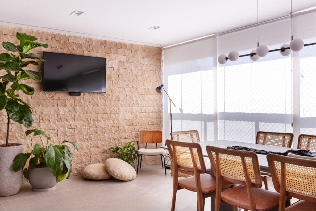 Sala de jantar com uma parede com pedra natural marrom e, a frente dela, uma mesa de madeira com cadeiras e uma janela com cortinas ao lado