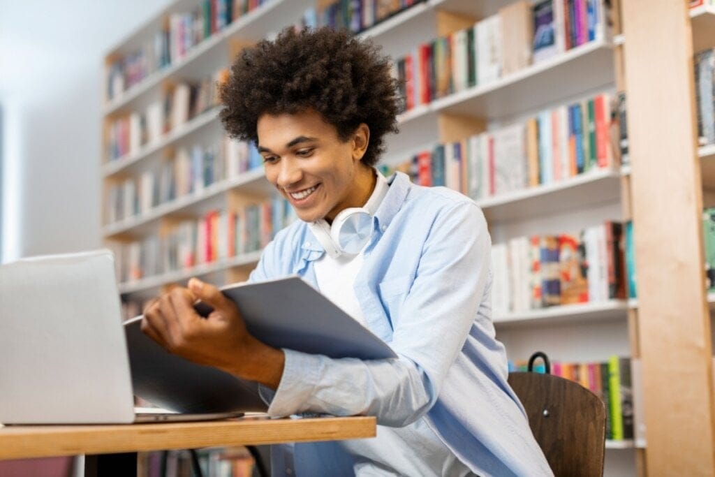Jovem com cabelo cacheado, usando camiseta branca e camisa azul com um fone de ouvido no pescoço, estudando em biblioteca