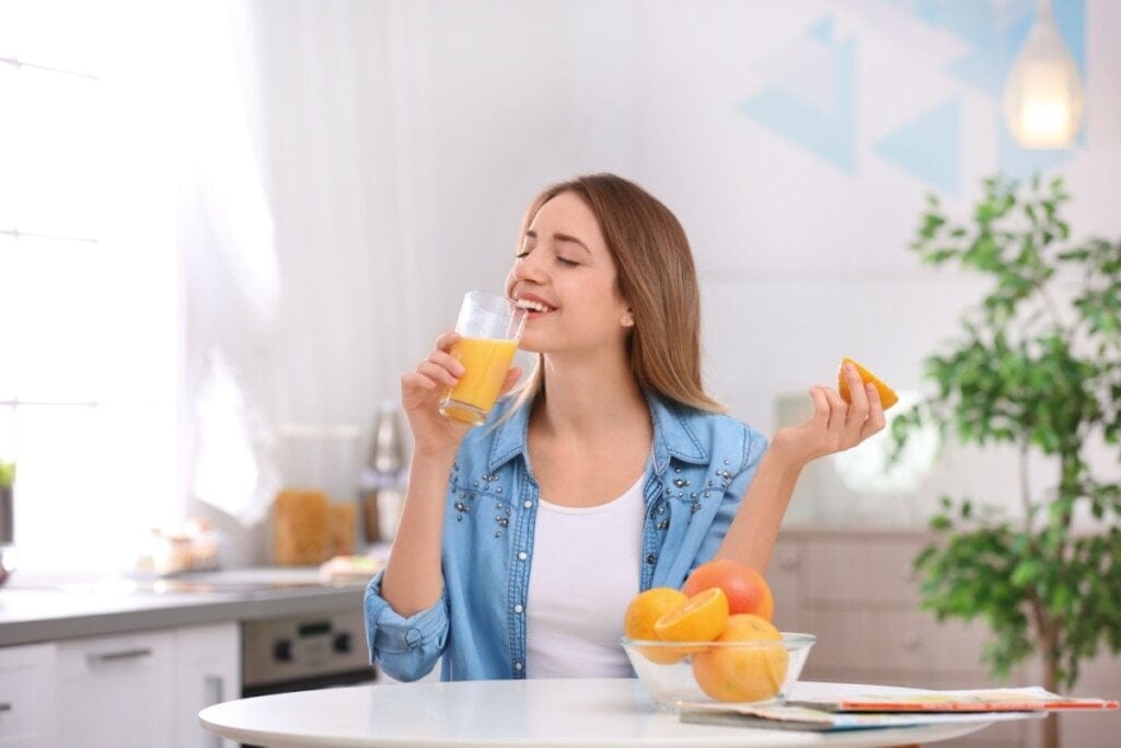 Mulher sentada em bancada tomando suco de laranja, ela está vestindo uma camiseta branca e uma camisa azul.