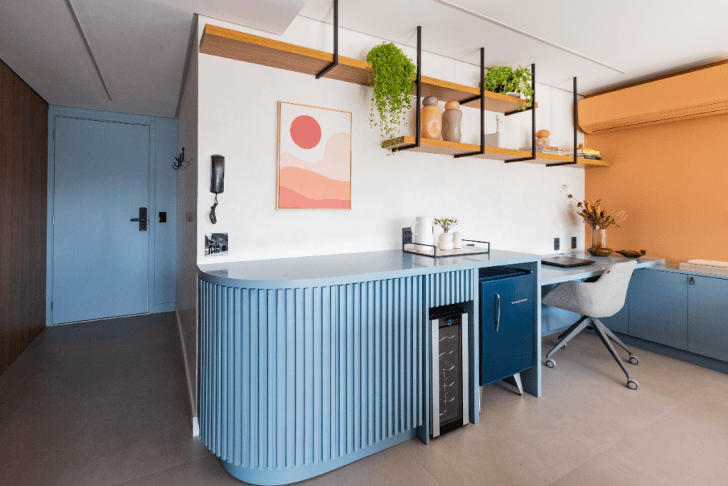 imagem de bar em casa com mesa e geladeira pequena em tons de azul, parede branca com prateleiras e planta e cadeira cinza ao fundo em home office