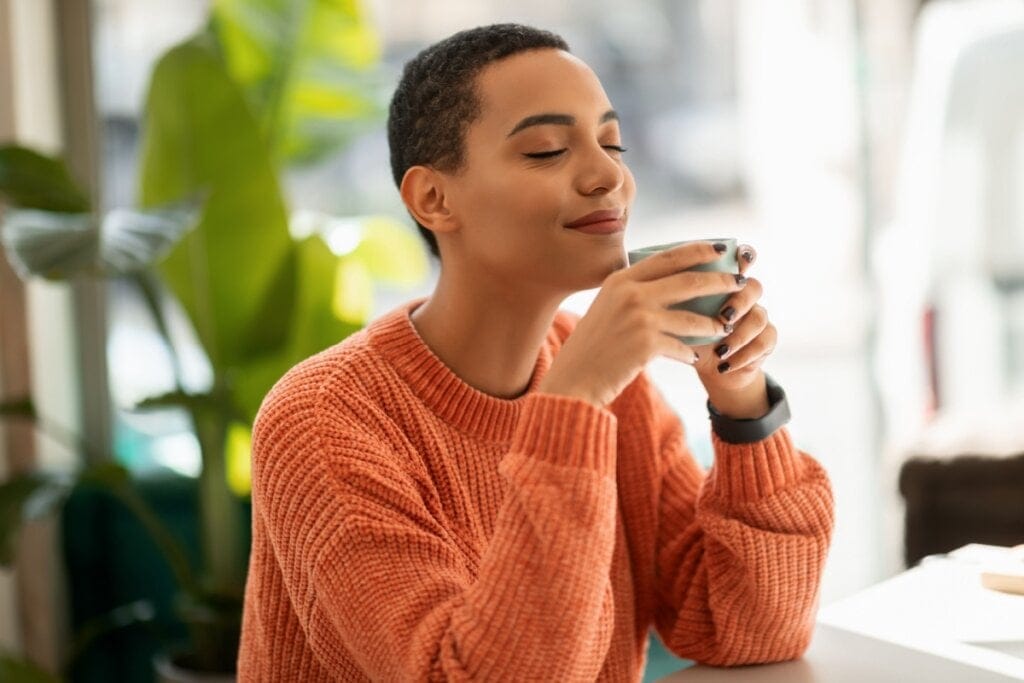 Jovem com o cabelo curto, usando um suéter laranja com uma xícara verde na mão tomando chá