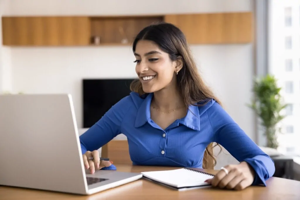 mulher camisa azul, sorrindo e sentada digitando em notebook em mesa