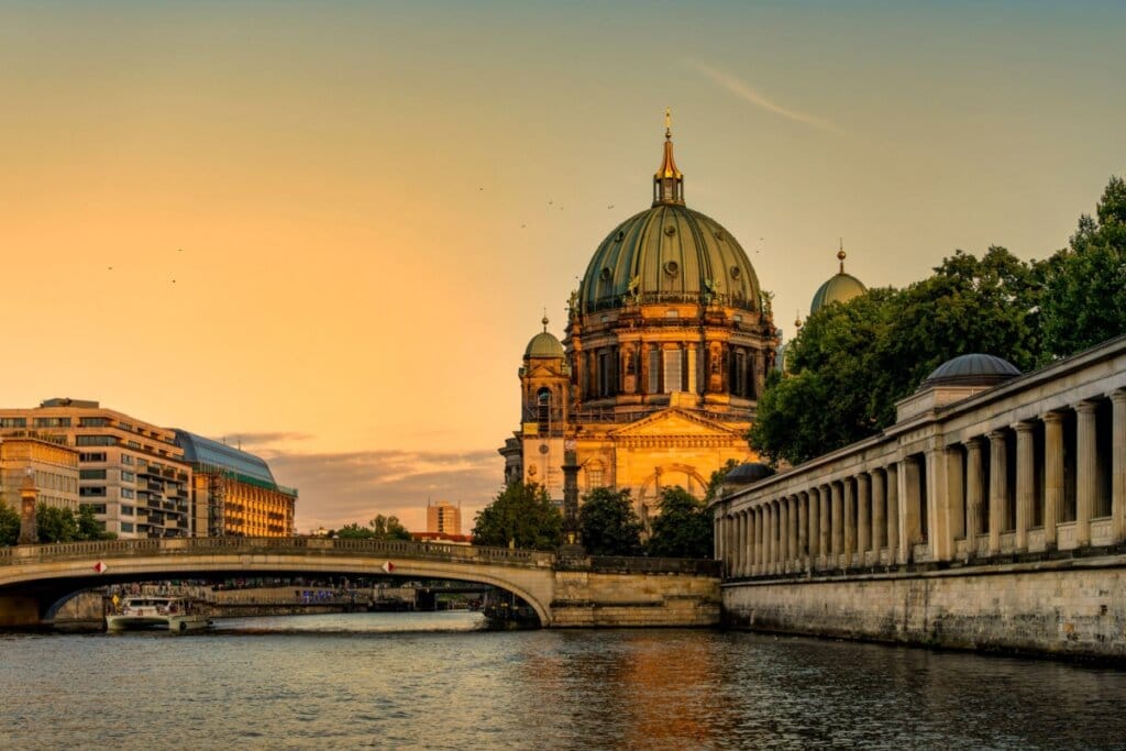 Imagem de uma construção neoclássica em frente a uma ponte e um rio abaixo dela ao pôr do sol em Berlim