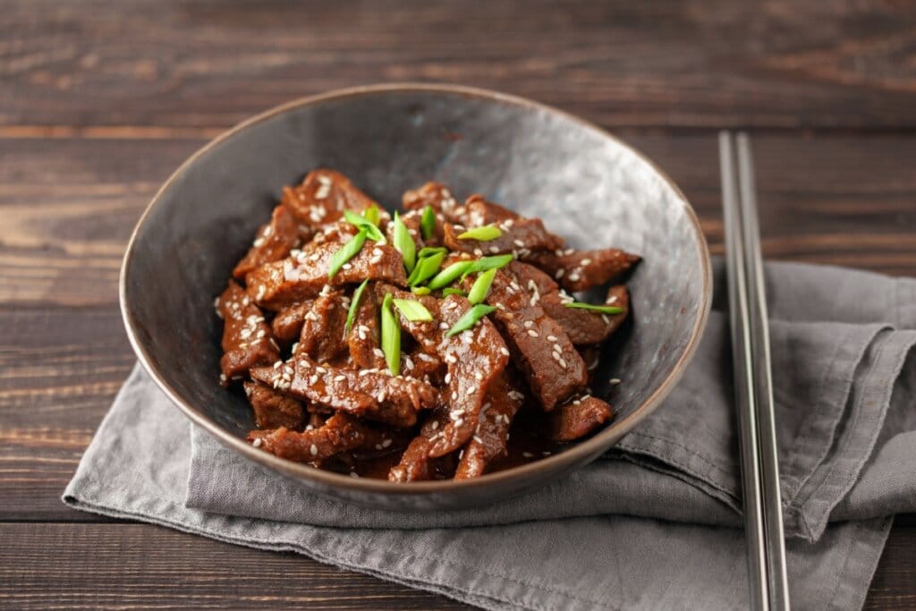 Tiras de carne marinadas e grelhadas servidas com gergelim e cebolinha em tigela de cerâmica cinza em cima de guardanapo de pano cinza com hashis ao lado em mesa de madeira 