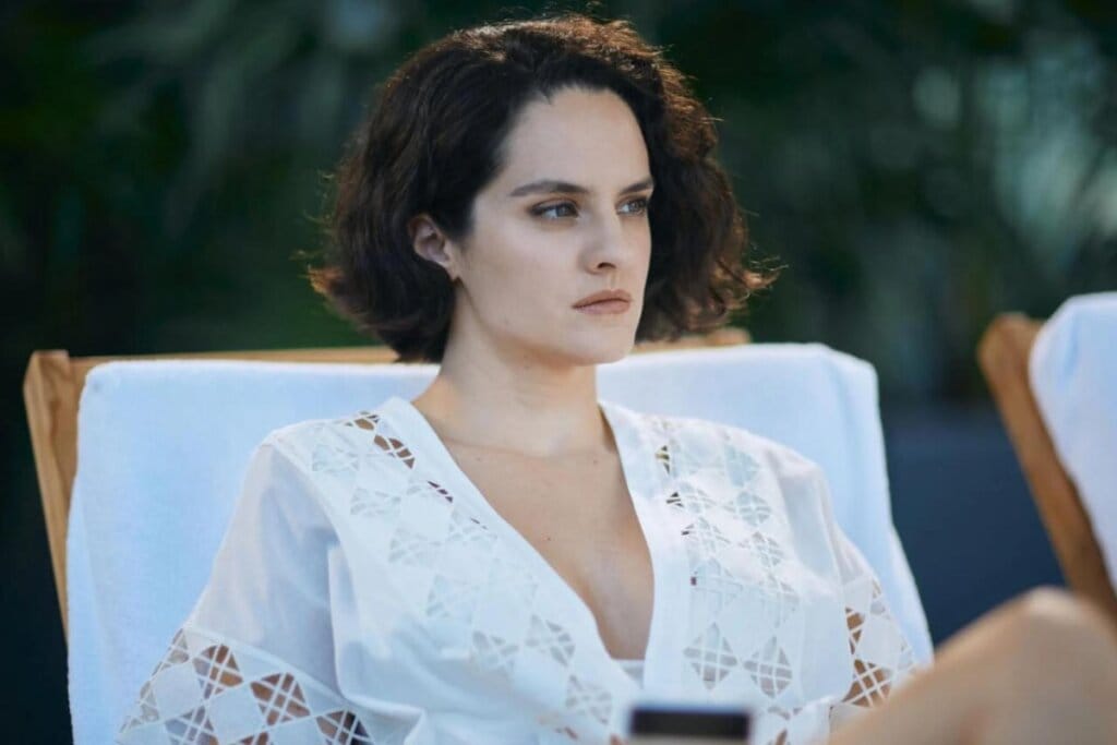 Cena do filme "Emmanuelle" com a protagonista sentada olhando para a frente e usando um vestido branco 