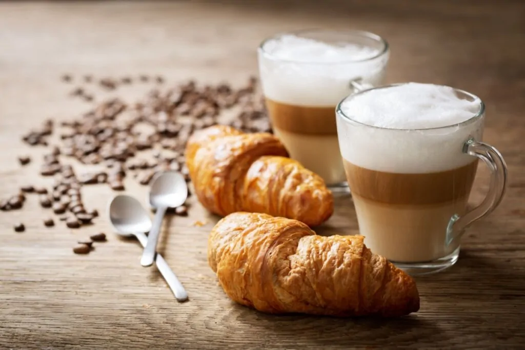 Duas canecas transparentes com Latte Macchiato em uma mesa de madeira e ao lado de dois croissant