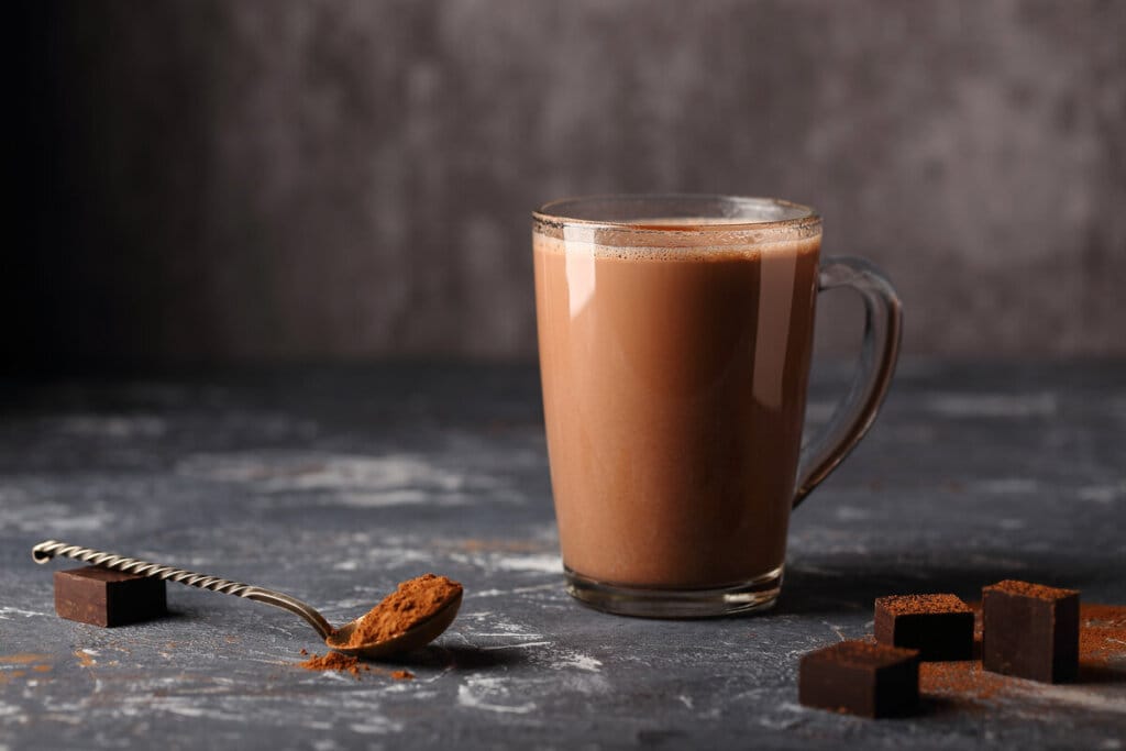 Caneca de vidro transparente com chocolate quente. colher com chocolate em pó e cubos de chocolate ao lado