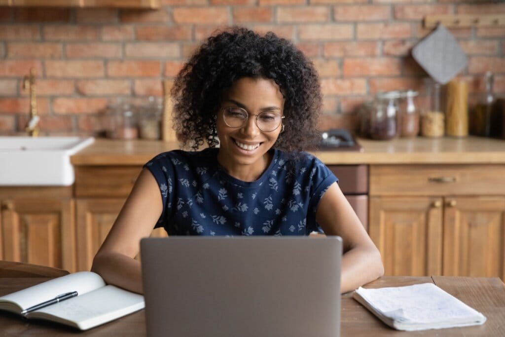 Jovem com o cabelo cacheado curto, com camiseta azul estampada escrevendo no notebook