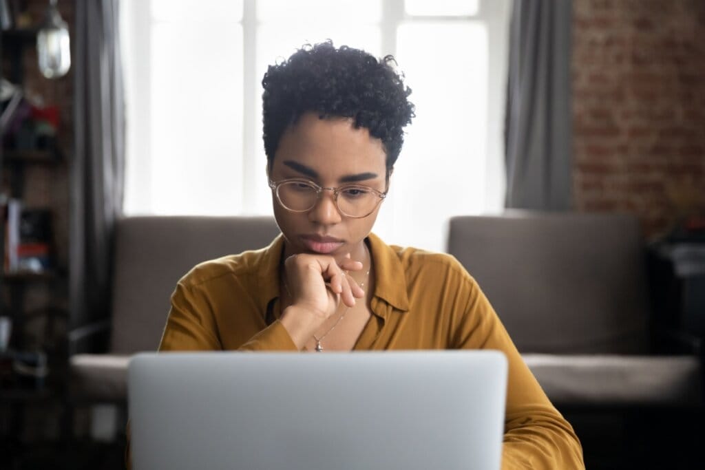 Jovem com o cabelo cacheado curto, usando óculos marrom e camisa marrom com a mão no queixo enquanto escreve no notebook