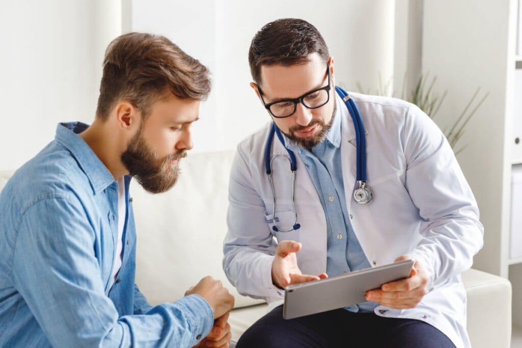 Homem e médico sentados conversando. Médico mostrando algo em uma prancheta 