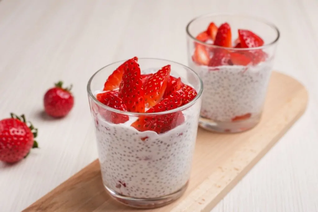 Dois copos com pudim de chia com linhaça e com morangos cortados em cima do pudim. Em cima de uma bandeja na bancada e morangos ao lado