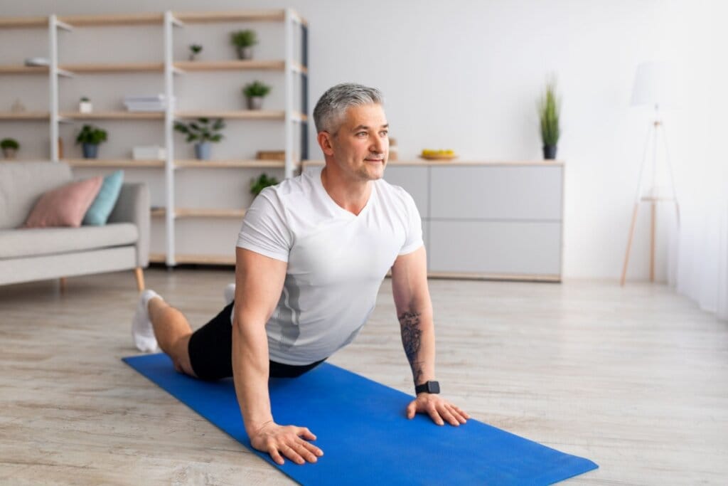 Homem de cabelos grisalhos praticando yoga em casa enquanto realiza a postura da cobra sobre um tapete azul