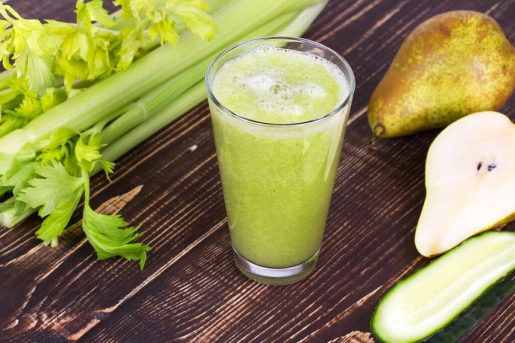 Copo de vidro com suco verde em cima de uma mesa de madeira com aipo, pera e pepino