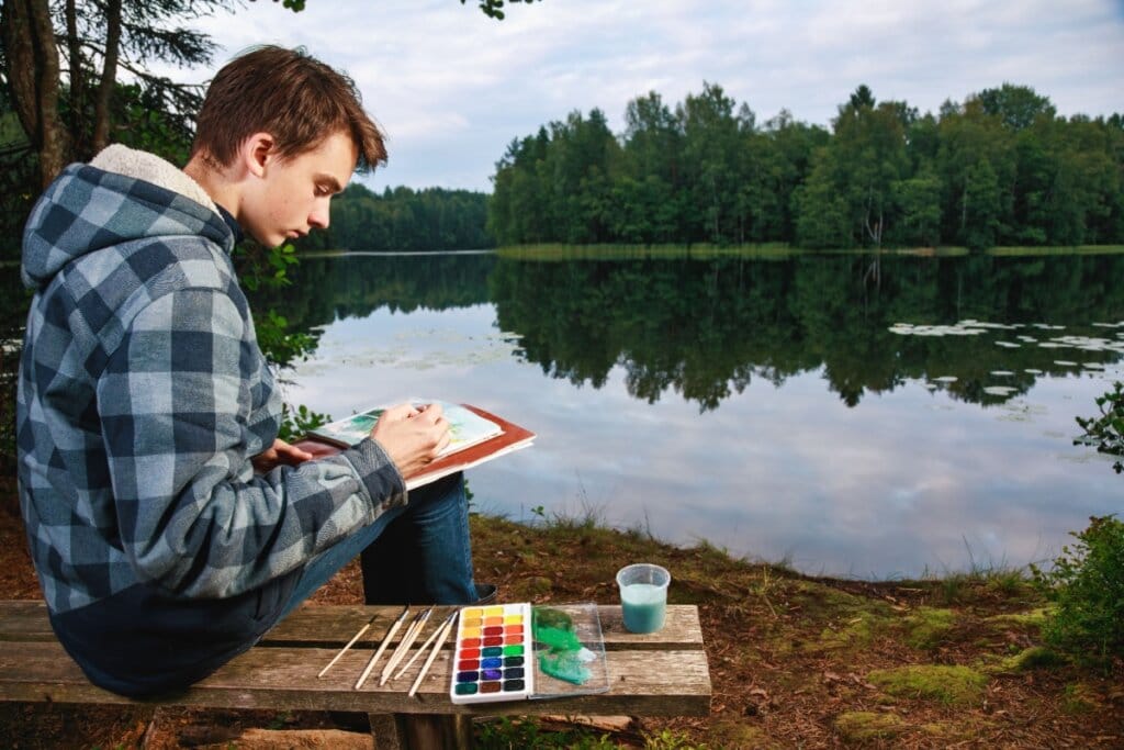 Adolescente sentado à beira de um lago pintando com aquarela em um momento de introspecção 