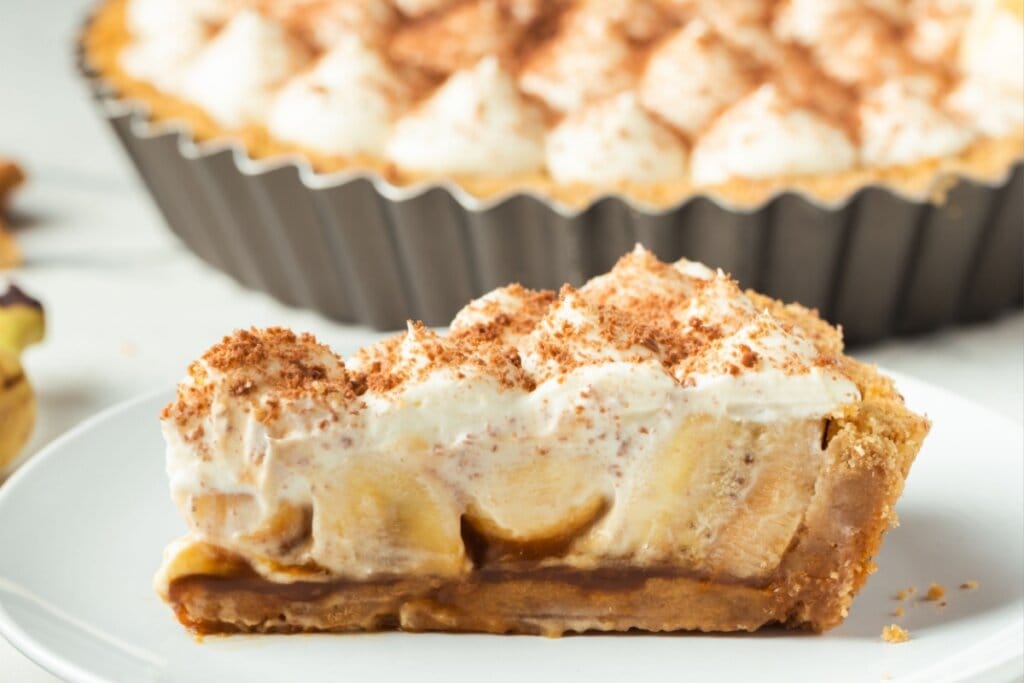 Pedaço de banoffee servido em prato branco em cima de mesa branca