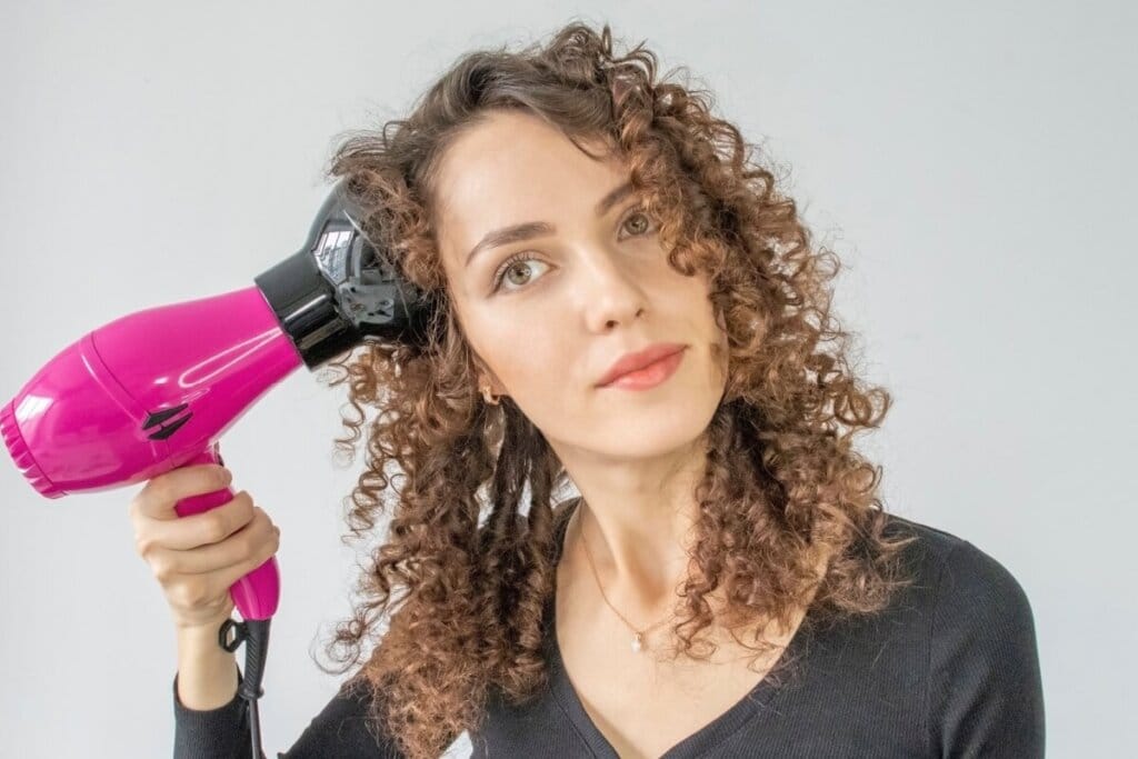 Mulher com o cabelo cacheado claro, usando secador com difusor para secar o cabelo e em fundo cinza