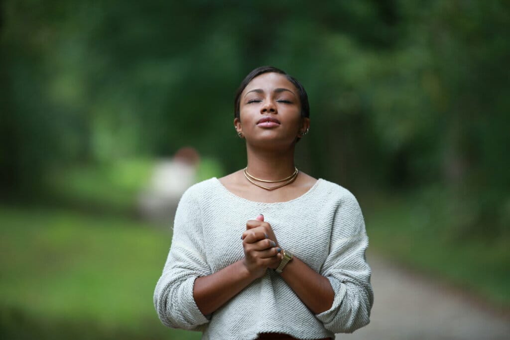 Uma mulher de olhos fechados vestindo blusa branca em um parque com as mãos juntas 