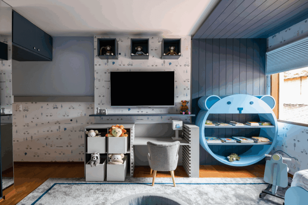 Quarto infantil com papel de parede azul e branco estampado, com nichos e TV na parede, com uma escrivaninha com cadeira cinza e ao lado uma prateleira de livros azul em forma de ursinho