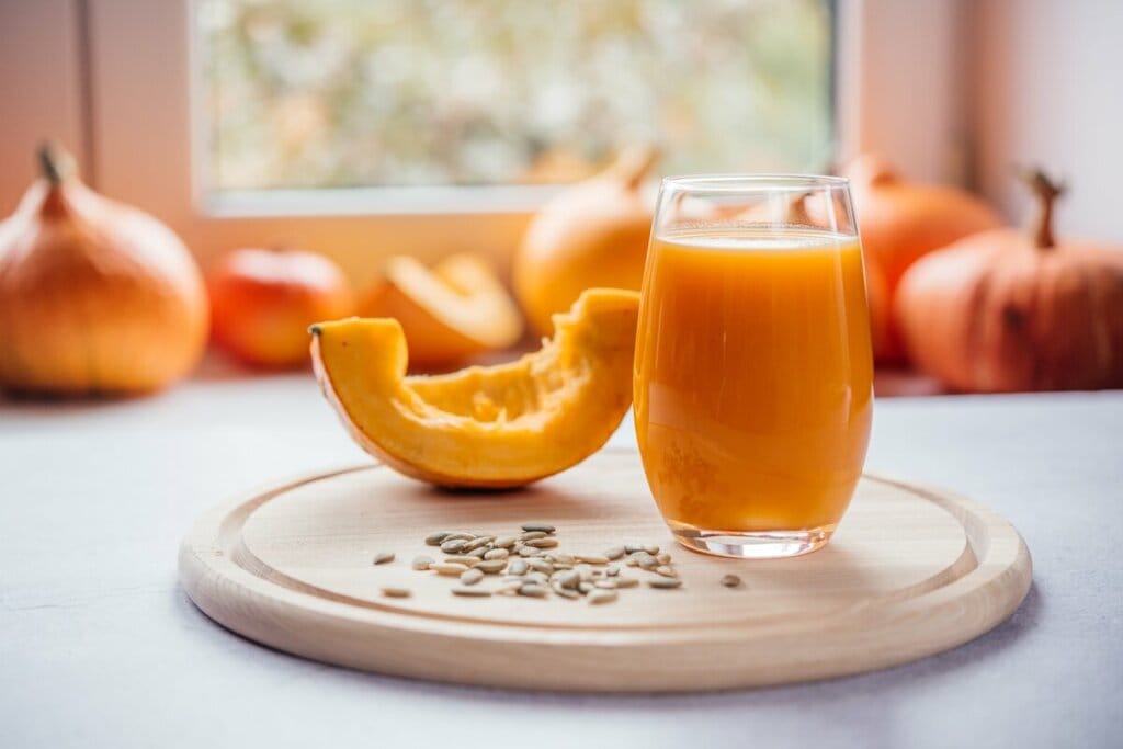 Copo de vidro com suco de abóbora e um pedaço de abóbora em cima de uma tábua de madeira