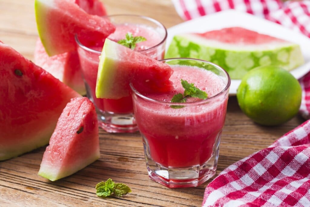 Copo de vidro com suco de melancia, pedaço de melancia na borda do copo e melancia e limão ao lado 