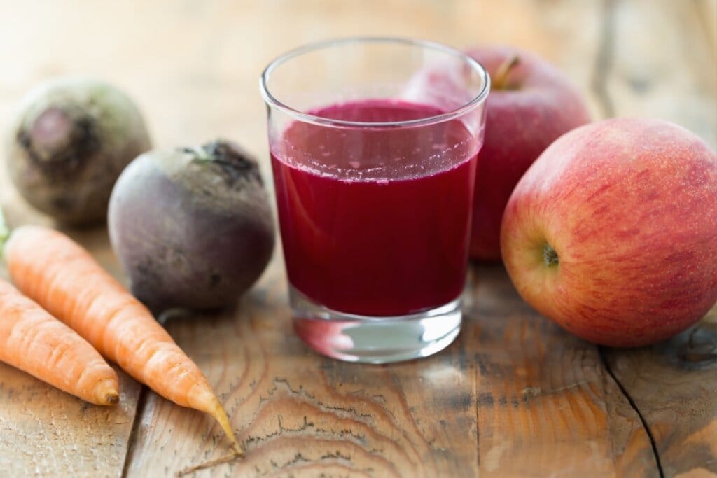 Suco de beterraba, maçã e cenoura servido em copo de vidro com beterraba, maçã e cenoura ao lado