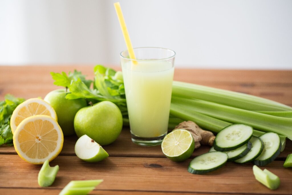 Copo de suco verde detox acompanhado de maçã-verde, pepino, aipo, limão, gengibre e folhas frescas sobre mesa de madeira
