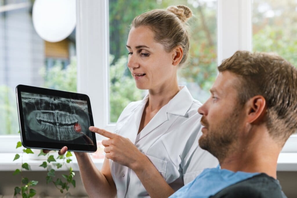 Dentista mostrando em um tablet a radiografia da boca de um paciente, apontando o dente do siso que passará por cirurgia de extração