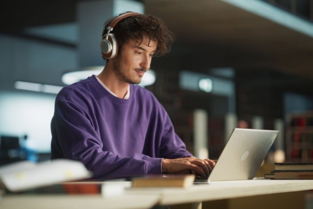Jovem com o cabelo curto e usando blusa de moletom roxa, e fones de ouvido prata, estudando no notebook