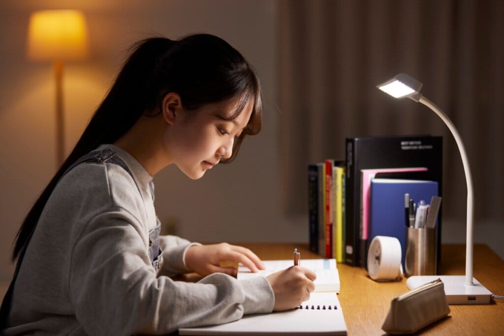 Menina de cabelo preto preso, blusa cinza e escrevendo em caderno em cima de mesa com luminária