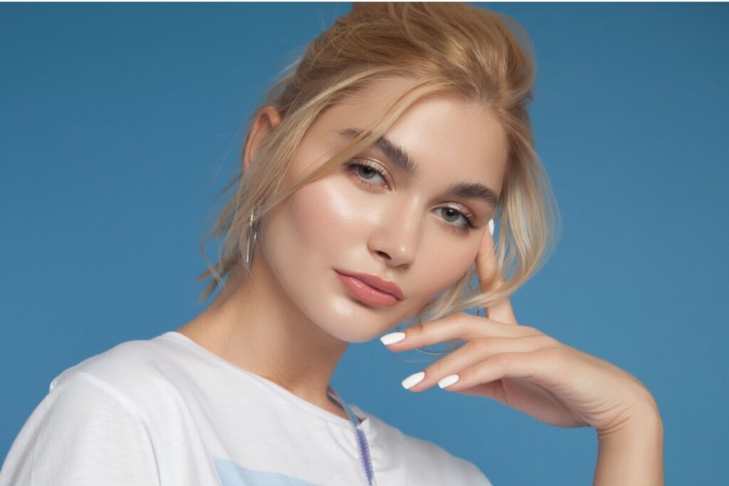 Mulher com o cabelo loiro, preso com fios soltos e com a mão apoiada no rosto, usando camiseta branca em fundo azul com a pele com tons naturais e saudaveis