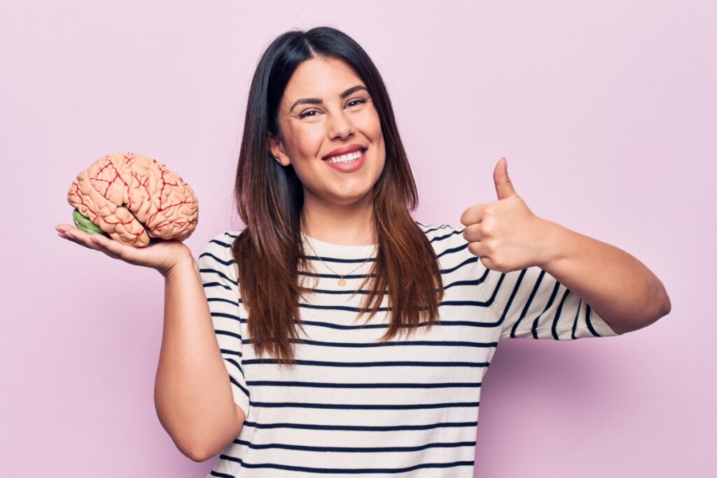 mulher de cabelo longo, sorrindo, fazendo "joia" com o dedo e segurando cérebro na mão