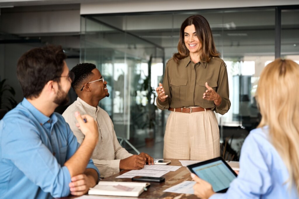 Mulher conversando com equipe de trabalho, ela tem o cabelo curto castanho e está usando uma camisa de botões verde e calça bege