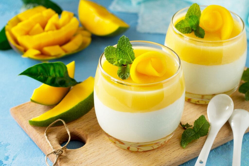 Pana cotta de manga servido em copos de vidro em cima de tábua de madeira com manga fatiada ao redor