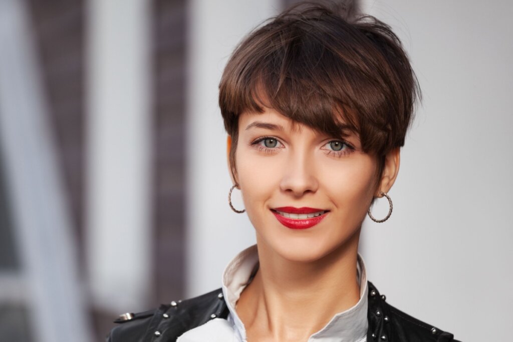 Mulher com o cabelo curto, liso e franja, usando camisa de botão e jaqueta de couro, brincos de argora e batom vermelho