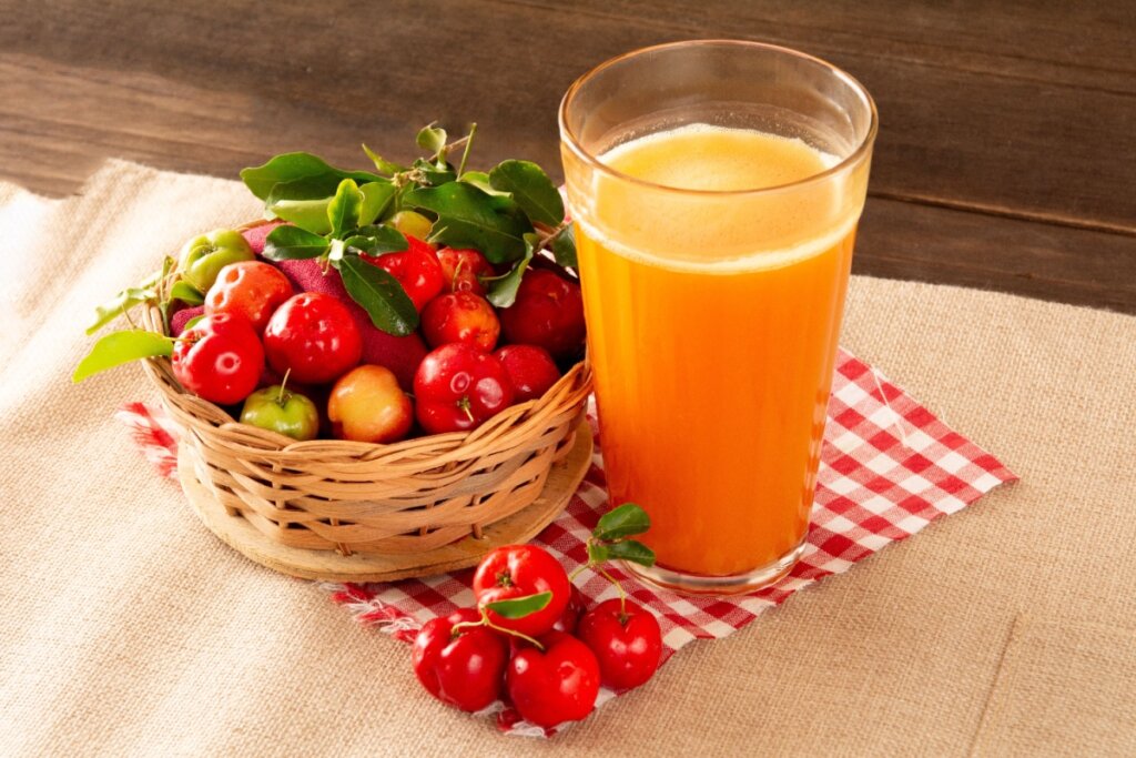 Suco de acerola ao lado de cestinha com acerolas frescas em cima de pano branco e vermelho
