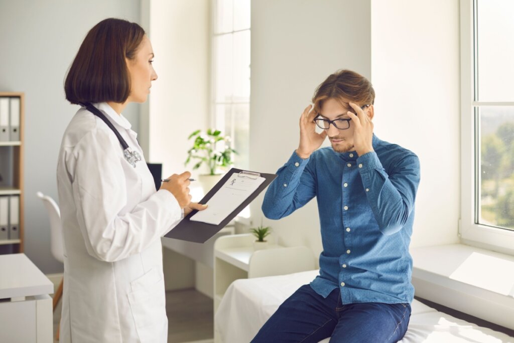 Médica em consultório avaliando paciente, que está com as mãos na cabeça, sugerindo enxaqueca