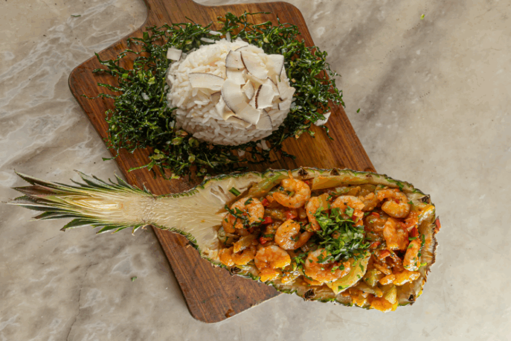 Ensopado de camarão no abacaxi com arroz de coco e couve crispy servido em tábua de madeira em cima de mesa de pedra